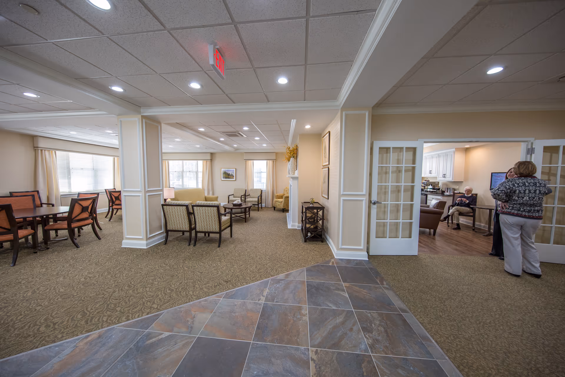 Interior view of a senior living community common area with carpeted and tiled flooring. The space includes a seating area with chairs and tables, and a dining area with multiple tables and chairs near large windows. To the right, there is a room with glass-paneled double doors where two women are standing and an elderly man is seated, with a kitchen area visible in the background.