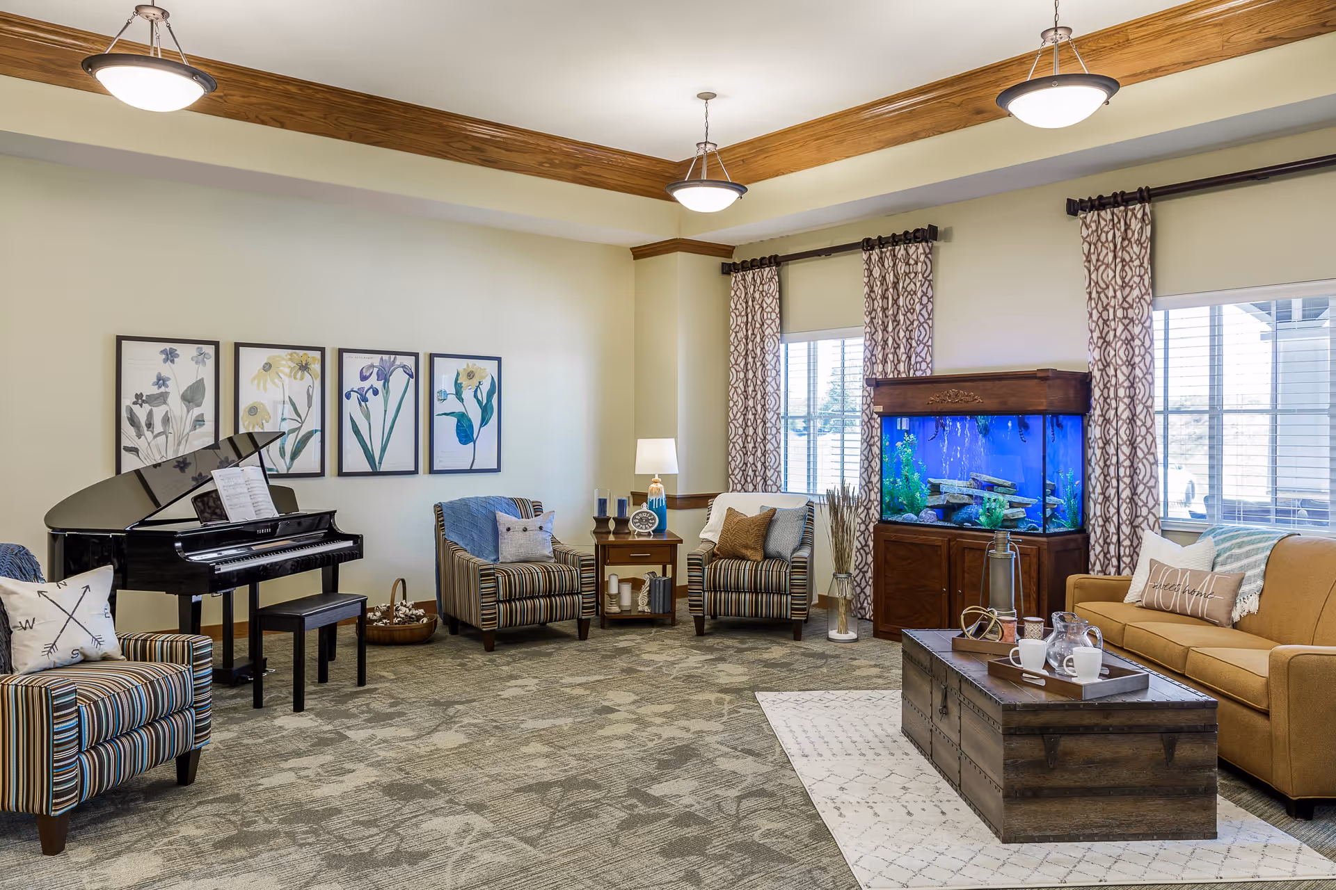 A cozy living room with a grand piano on the left, two striped armchairs with pillows, a wooden side table with a lamp and decorative items, a large fish tank between two windows with patterned curtains, and a mustard-colored sofa with pillows on the right. A wooden coffee table with a tray holding cups and a pitcher sits on a patterned rug in the center of the room. The ceiling has wooden beams and three hanging light fixtures.