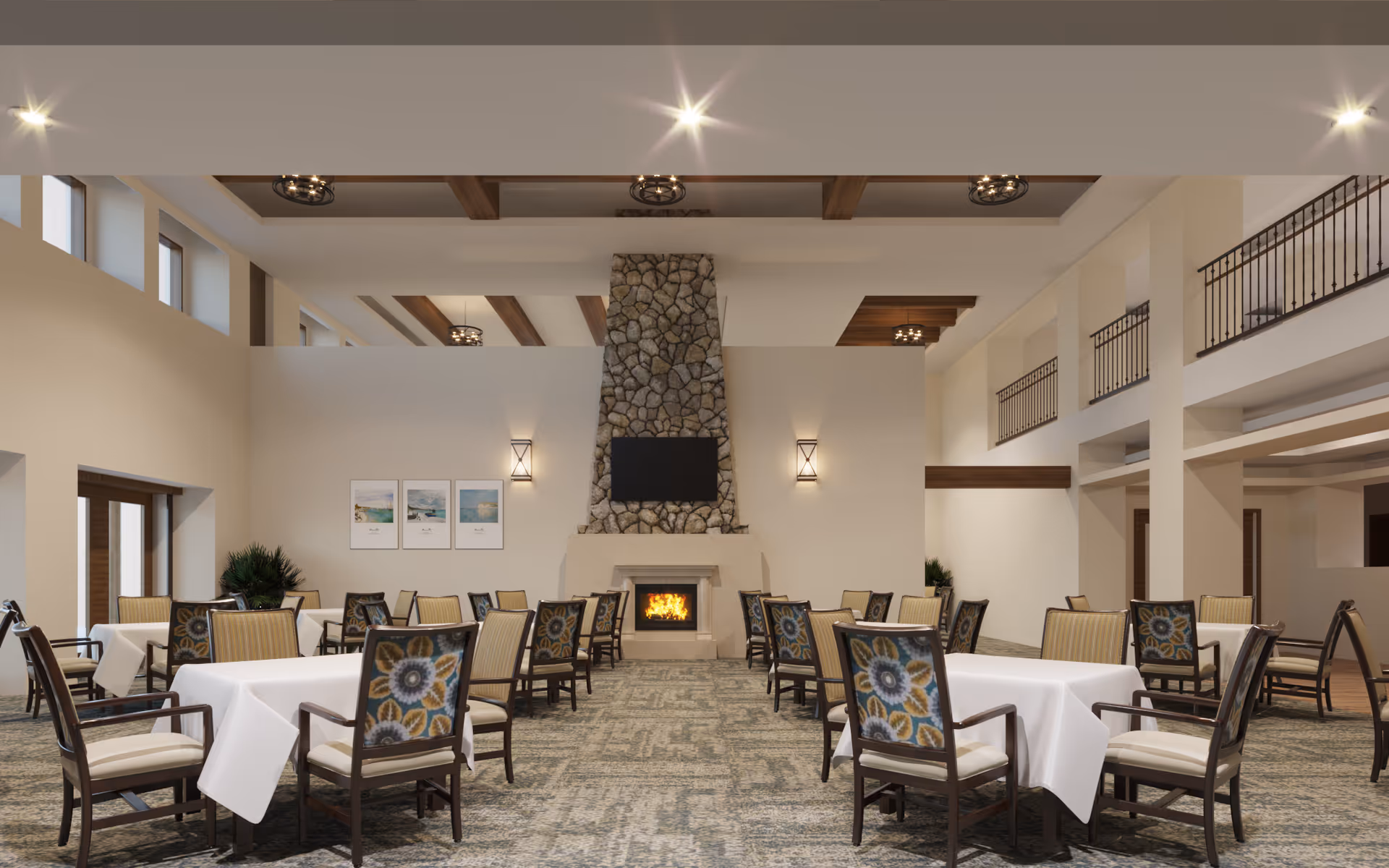A spacious dining room with multiple tables covered in white tablecloths and surrounded by chairs with floral patterned backs and cushioned seats. The room features a large stone fireplace with a fire burning and a flat-screen TV mounted above it. The ceiling has wooden beams and modern light fixtures, and there are windows near the ceiling letting in natural light. The walls are light-colored, and there are framed pictures on one wall.