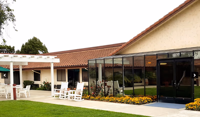 Outdoor patio area at Fountain Square Of Lompoc featuring white rocking chairs, tables with chairs under a pergola, a glass-enclosed sunroom, and a well-maintained lawn with flower beds.