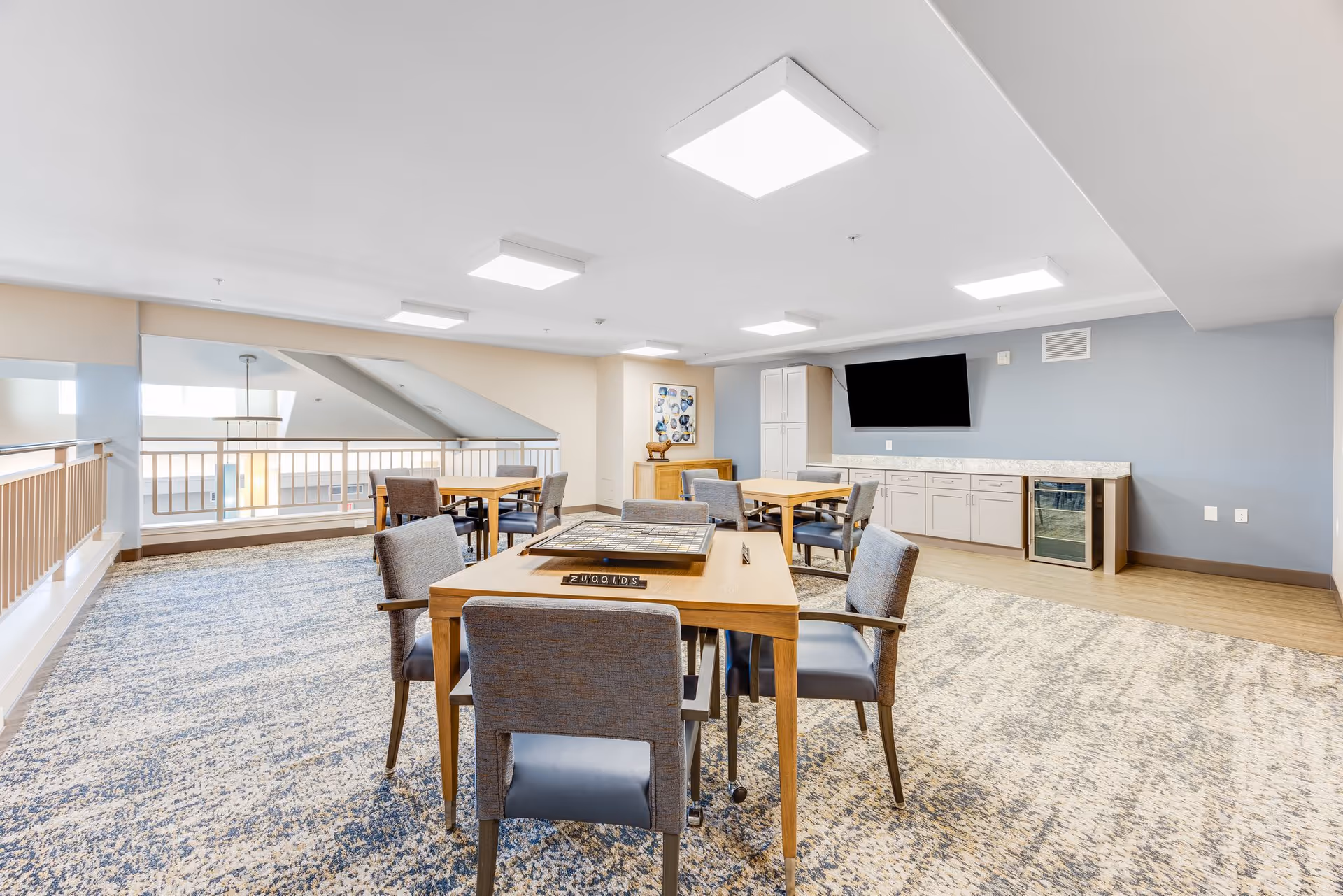 A bright, spacious common area with multiple tables and chairs arranged for group activities or socializing. The room features a patterned carpet, light-colored walls, a large flat-screen TV mounted on the wall, and a countertop with cabinets and a small refrigerator underneath. The area is well-lit with ceiling lights and has a railing overlooking a lower level.