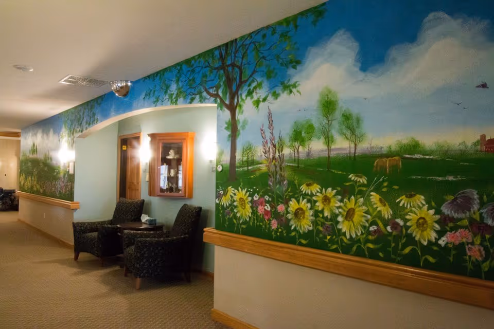Interior hallway of Prairie View Manor with a large mural on the wall depicting a scenic landscape with trees, flowers, and a blue sky with clouds. Two upholstered chairs and a small table are placed against the wall under an archway. The hallway is carpeted and softly lit with wall sconces.