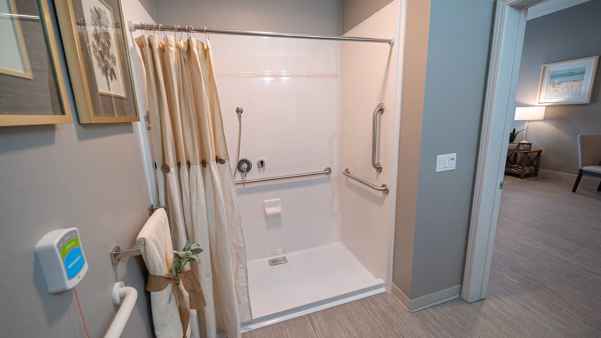 A clean and accessible shower area with beige and white shower curtains, grab bars on the walls, and a handheld showerhead. A towel with a decorative ribbon and greenery is hanging on a towel rack next to the shower. The shower is adjacent to a room with a chair, side table, lamp, and framed artwork on the wall.