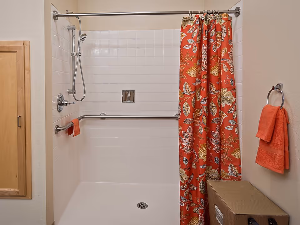 A bathroom shower area with white tiled walls and a silver handheld showerhead. There is a silver grab bar along the back wall and a red shower curtain with a leaf pattern partially drawn to the right. A red towel hangs on a ring on the right wall, and a wooden cabinet door is visible on the left wall.