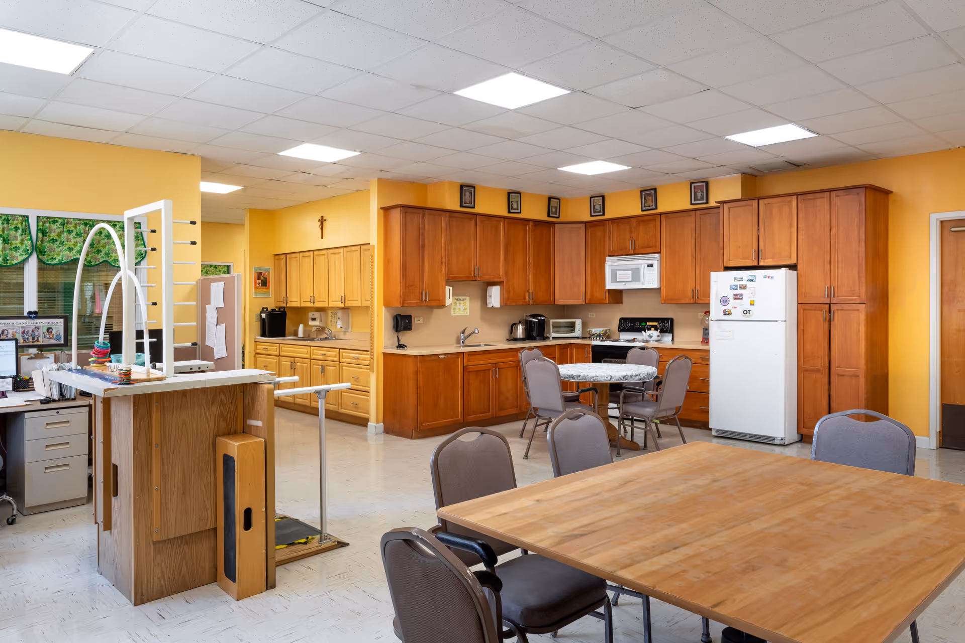 Communal dining and kitchenette area with tables, chairs, wooden cabinets, and a refrigerator in a senior living facility.