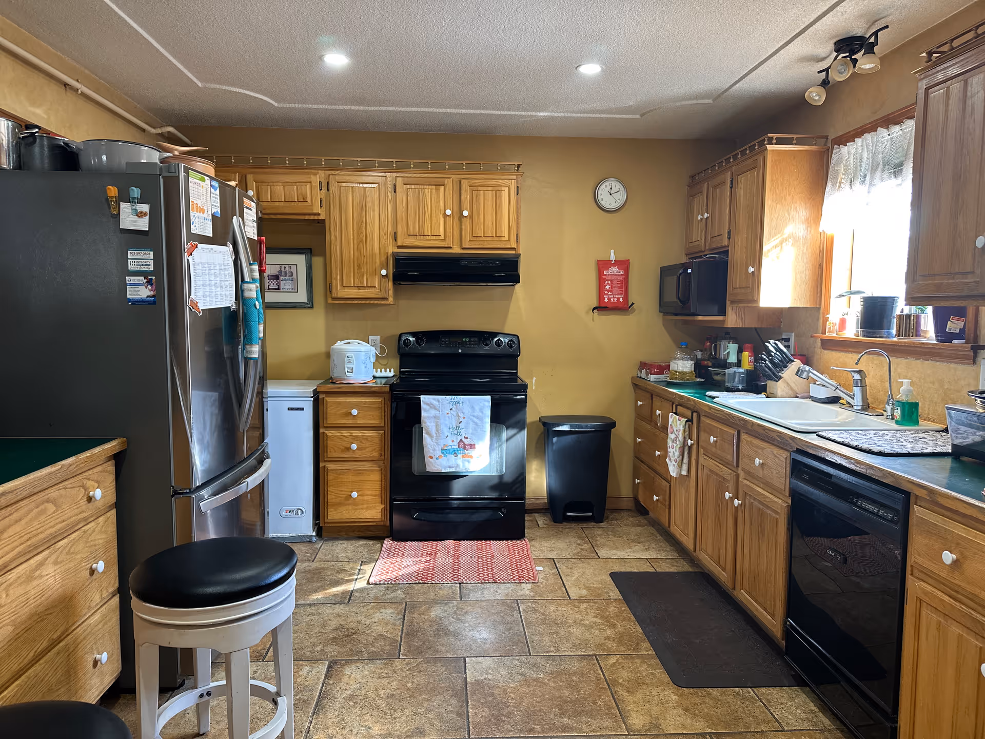 A kitchen with wooden cabinets, a black stove with a towel hanging on the handle, a black refrigerator with magnets and papers attached, a white stool with a black cushion, a dishwasher, a sink under a window with lace curtains, and various kitchen items on the counters.