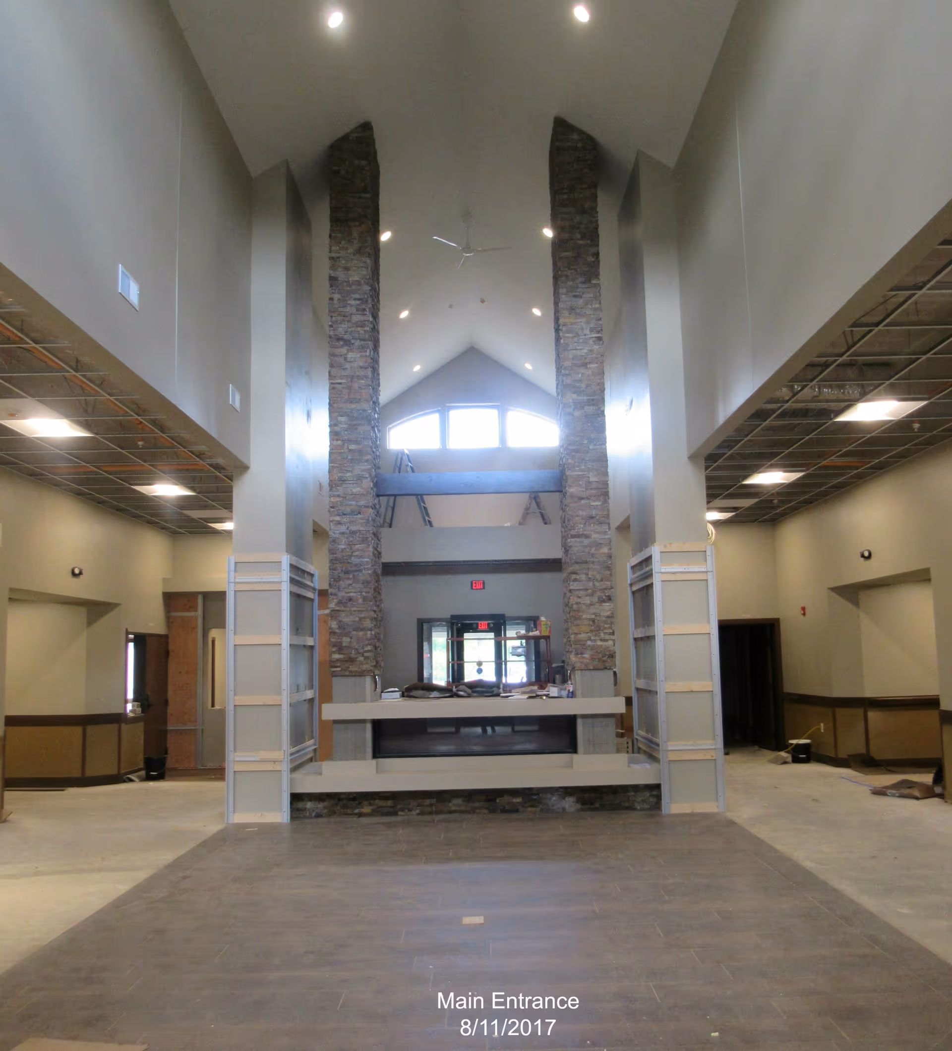 Interior view of a spacious main entrance area with high vaulted ceilings, two tall stone pillars framing a modern fireplace, and large windows letting in natural light. The floor is tiled, and the space appears to be under construction or renovation with some materials and equipment visible.