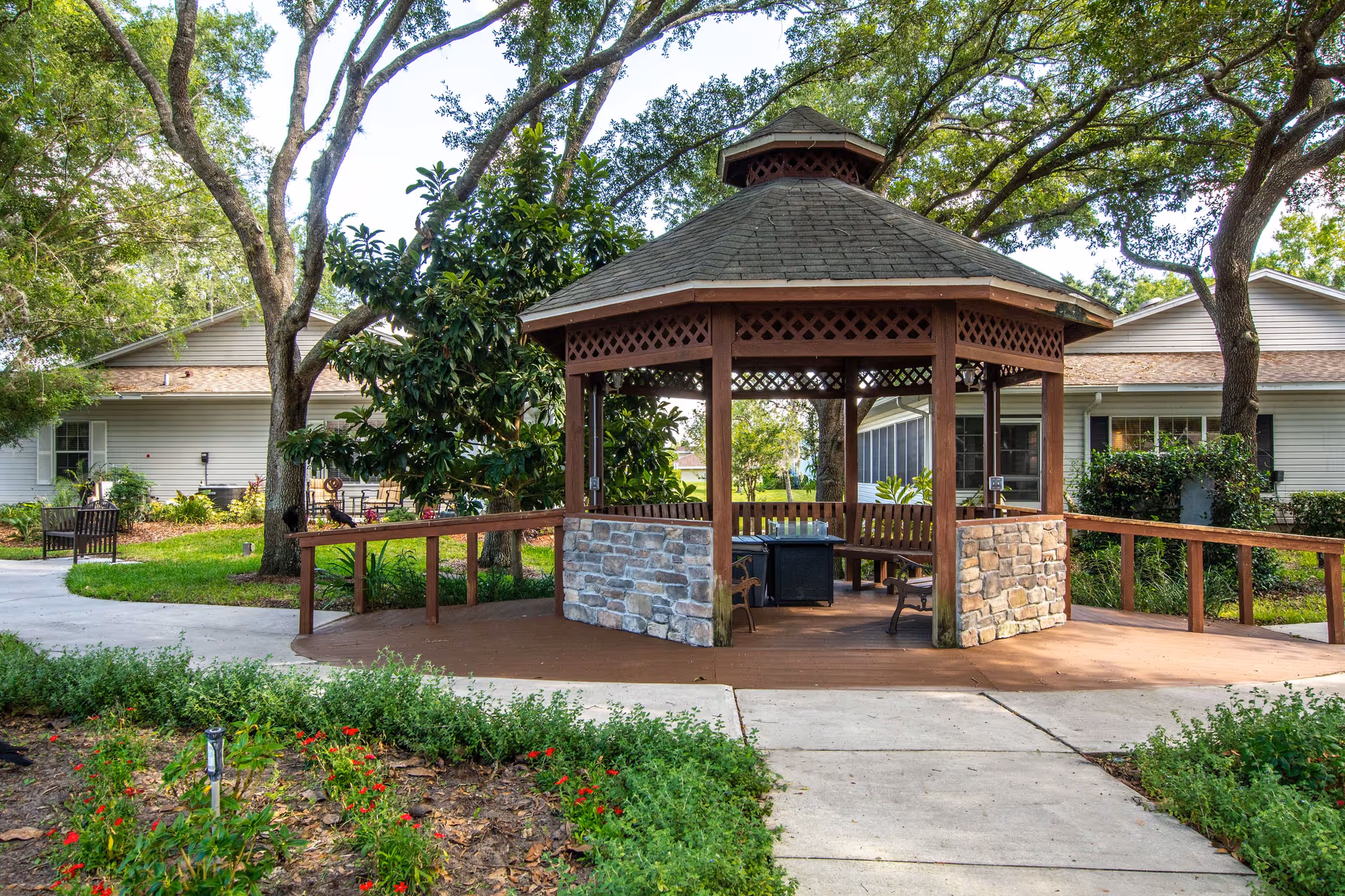 A wooden gazebo with stone accents on the lower half of its pillars, situated in a garden area with trees and greenery. There are benches inside the gazebo and a paved walkway leading to it. Residential buildings are visible in the background.