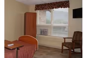 A simple senior living facility room with a single bed covered in a brown blanket, a wooden cabinet, a window with blinds and a valance, an air conditioning unit below the window, a striped armchair, and a small wall-mounted television.