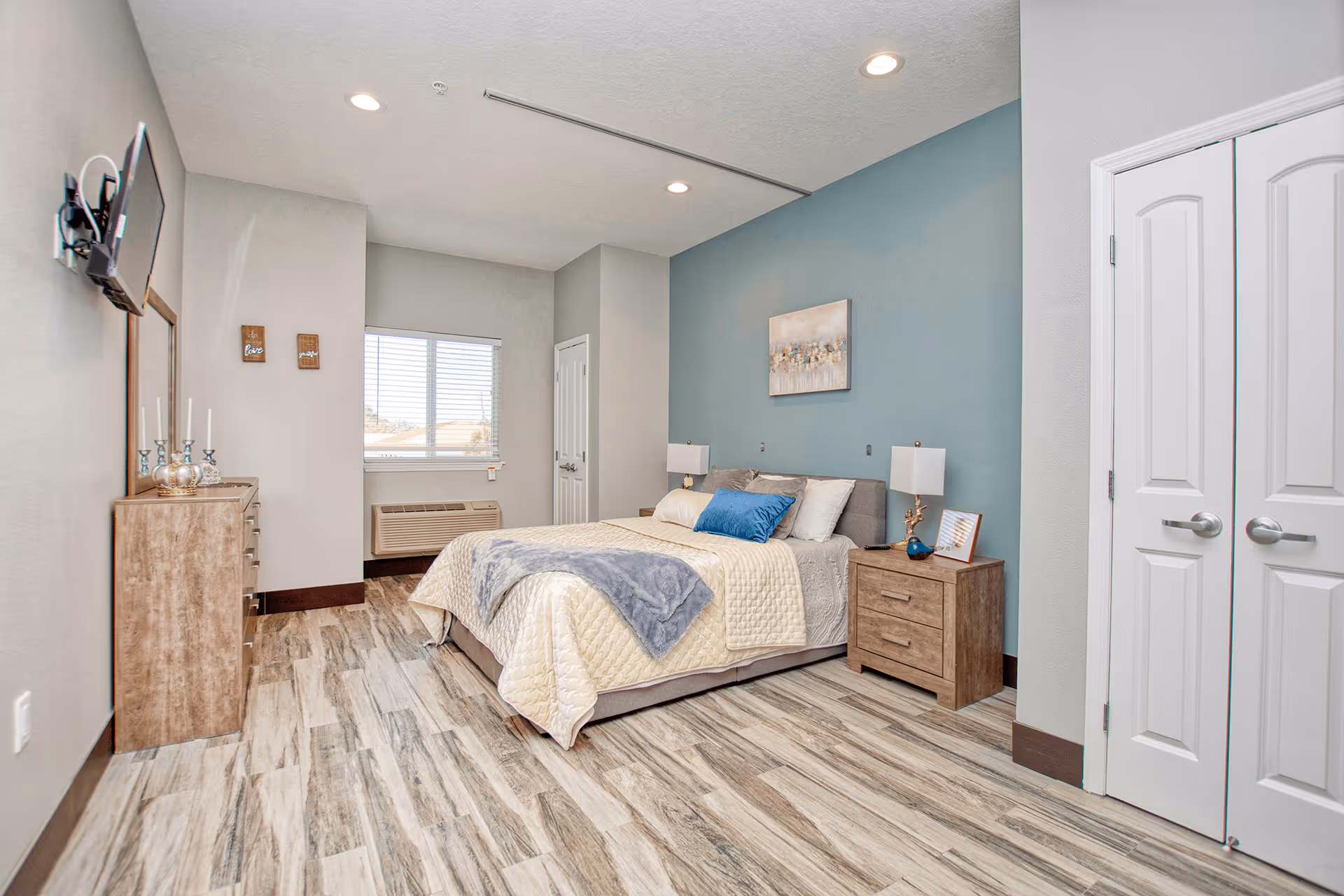 A bright and clean bedroom in an assisted living facility featuring a bed with beige and blue bedding, two wooden nightstands with lamps, a wooden dresser with decorative items, a wall-mounted TV, a window with blinds, and a closet with double doors. The room has light-colored walls with one accent wall painted blue and wood-patterned flooring.