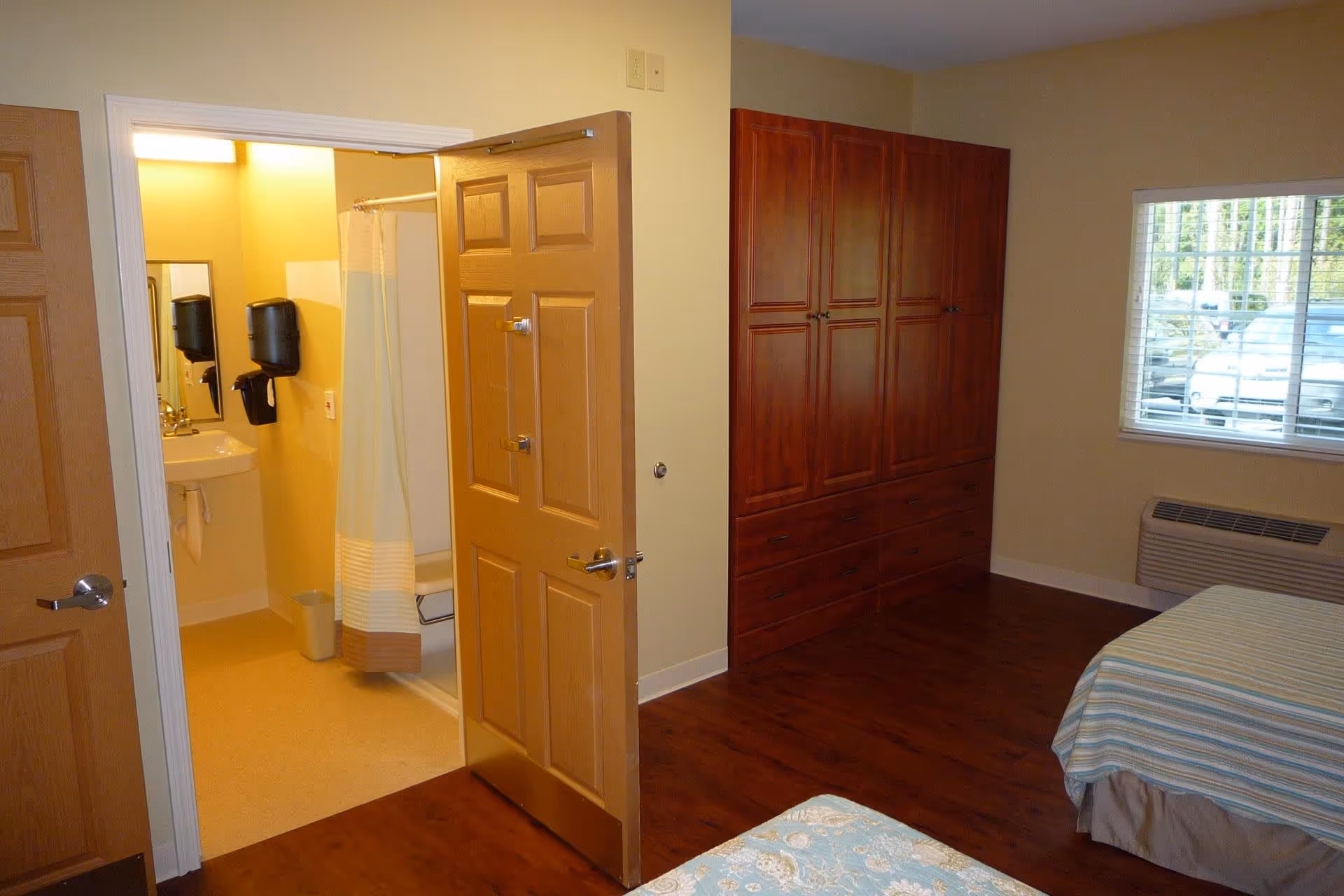 Interior view of a senior living facility room at New Hanover House showing part of a bedroom with two beds covered in patterned bedspreads, a large wooden wardrobe, a window with blinds, and an open door leading to a bathroom with a shower, sink, and hand sanitizer dispensers.