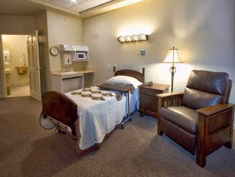 A senior living facility bedroom with a single bed covered in a white quilt, a wooden nightstand with a lamp, a brown leather recliner chair, and a small kitchenette area with a sink, microwave, and shelves. A bathroom is visible through an open door in the background.