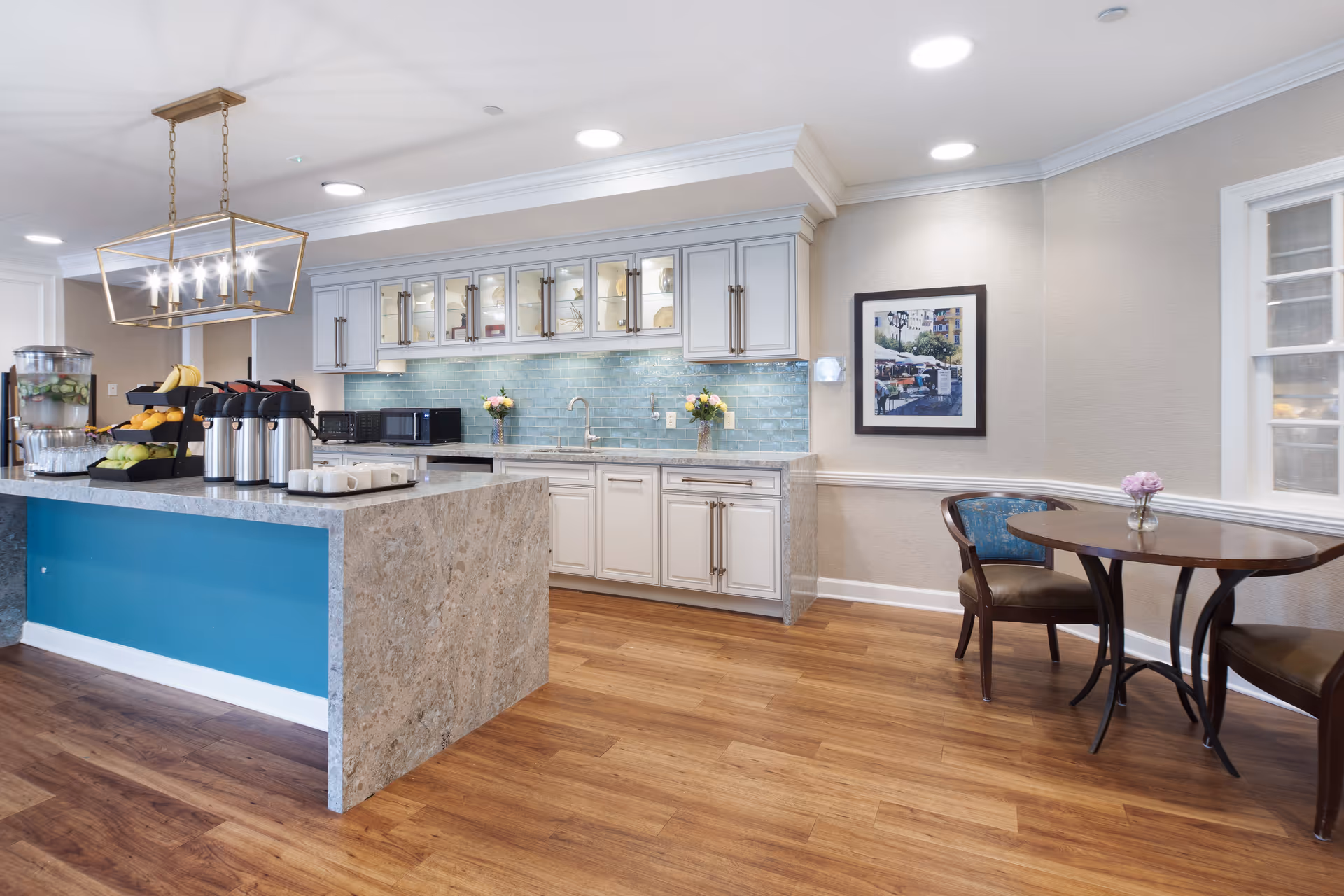A bright and modern kitchen area in a senior living facility with a large marble island featuring coffee dispensers, fresh fruit, and glassware. The back wall has white cabinets with glass doors, a blue tiled backsplash, and two vases with flowers. To the right, there is a small round wooden table with two chairs and a framed picture on the wall.