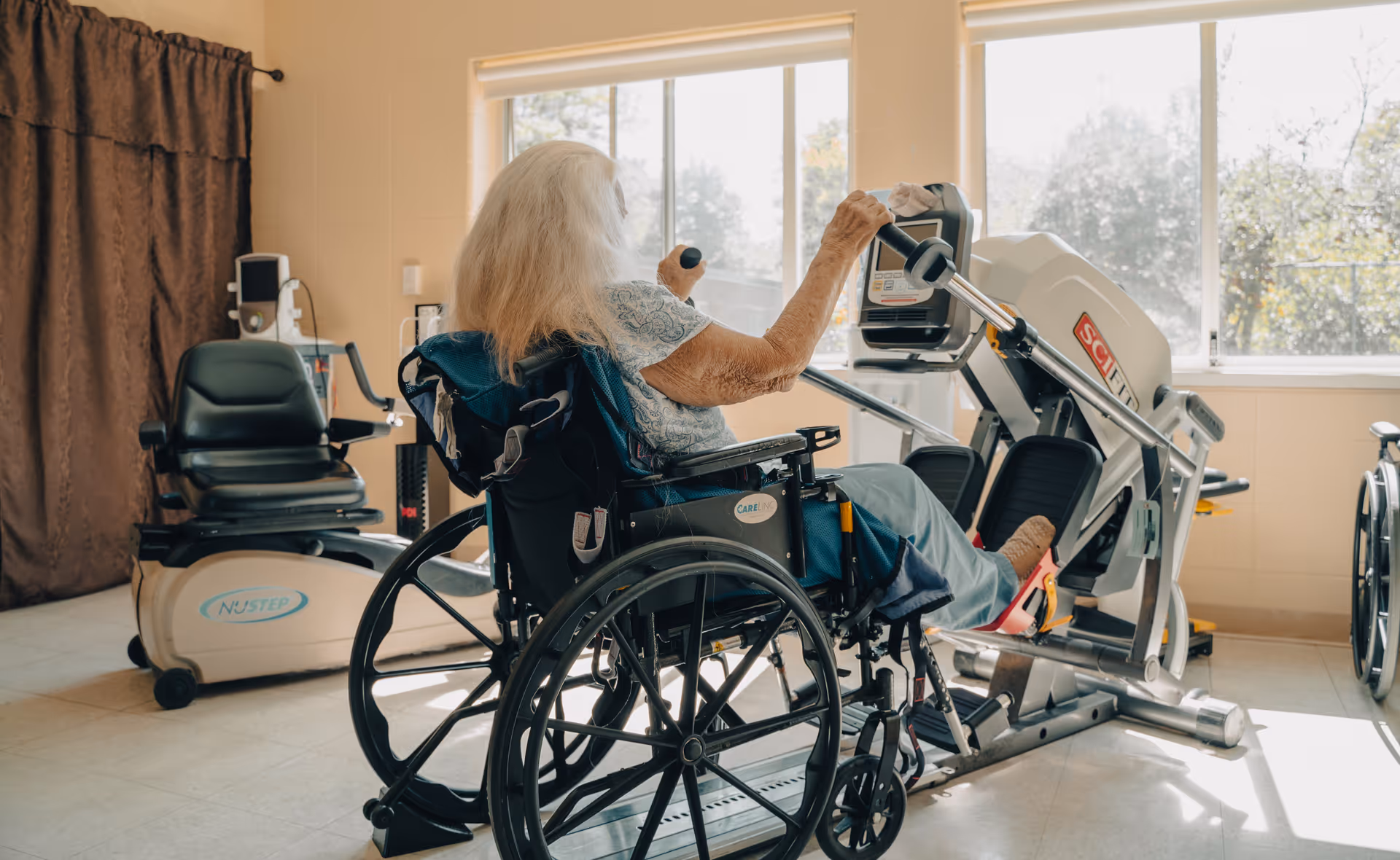 An elderly woman with long white hair in a wheelchair using a leg exercise machine in a well-lit room with large windows and exercise equipment.