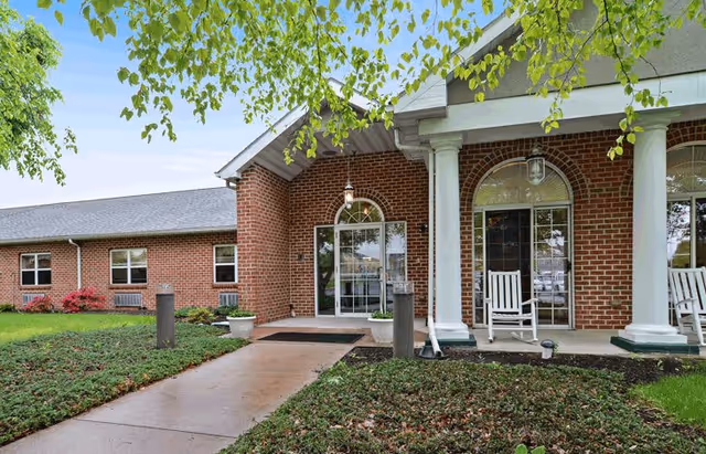 Front entrance of a brick building with white columns and rocking chairs on the porch, surrounded by green shrubs and trees under a clear blue sky.