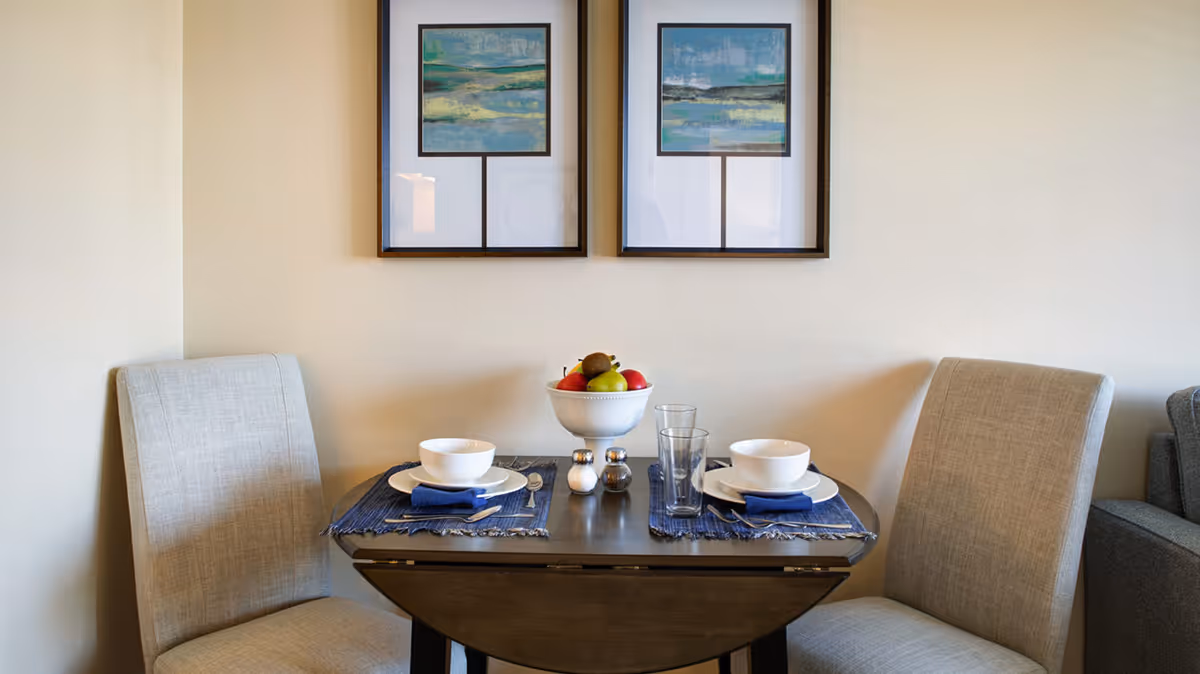 A small dining table set for two with white bowls, plates, blue napkins, silverware, and two glasses. A white bowl filled with assorted fruit is in the center of the table. Two beige upholstered chairs are positioned on either side of the table. Two framed abstract landscape paintings hang on the wall above the table.
