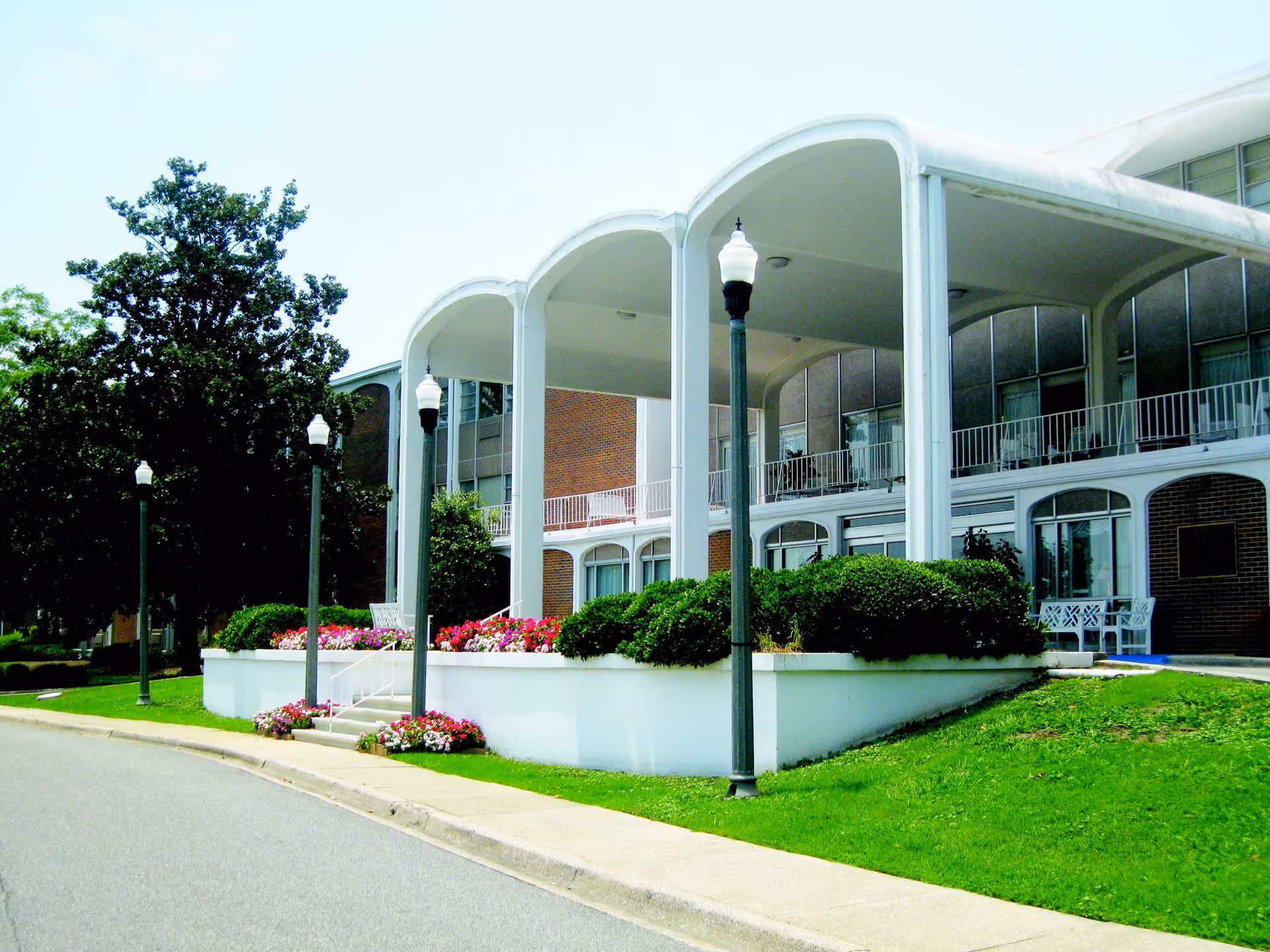 Exterior view of a senior living facility with a covered entrance supported by white columns, surrounded by green bushes and colorful flowers. There are several street lamps along the driveway and a large tree to the left.