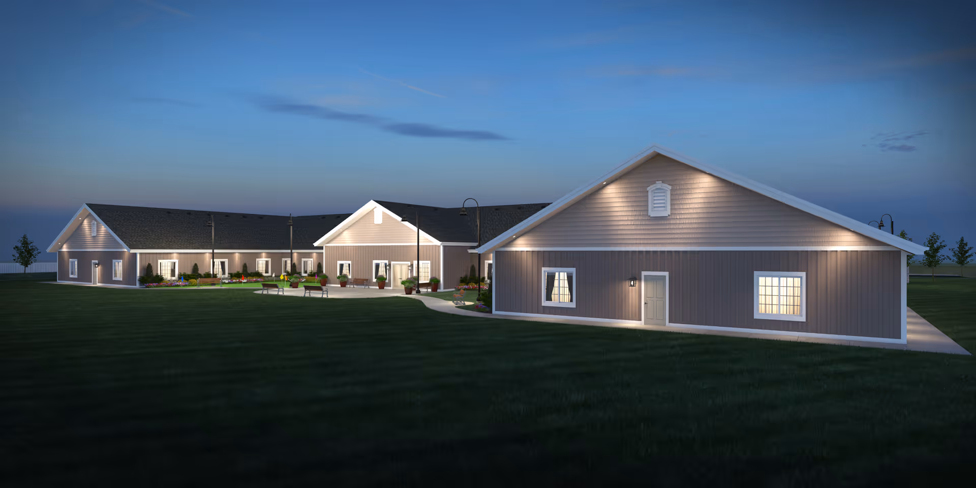 Exterior view of a single-story assisted living facility building at dusk with lights illuminating the entrance and windows, surrounded by a well-maintained lawn and a few trees in the background.