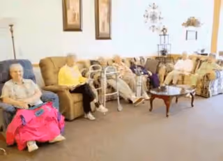 A group of elderly individuals sitting on couches and chairs in a well-lit common area with a coffee table in front of them. The room has framed artwork on the wall and a floor lamp in the corner.