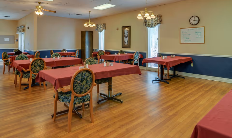 Dining room with multiple tables covered in red tablecloths and floral-upholstered chairs on a wood floor.