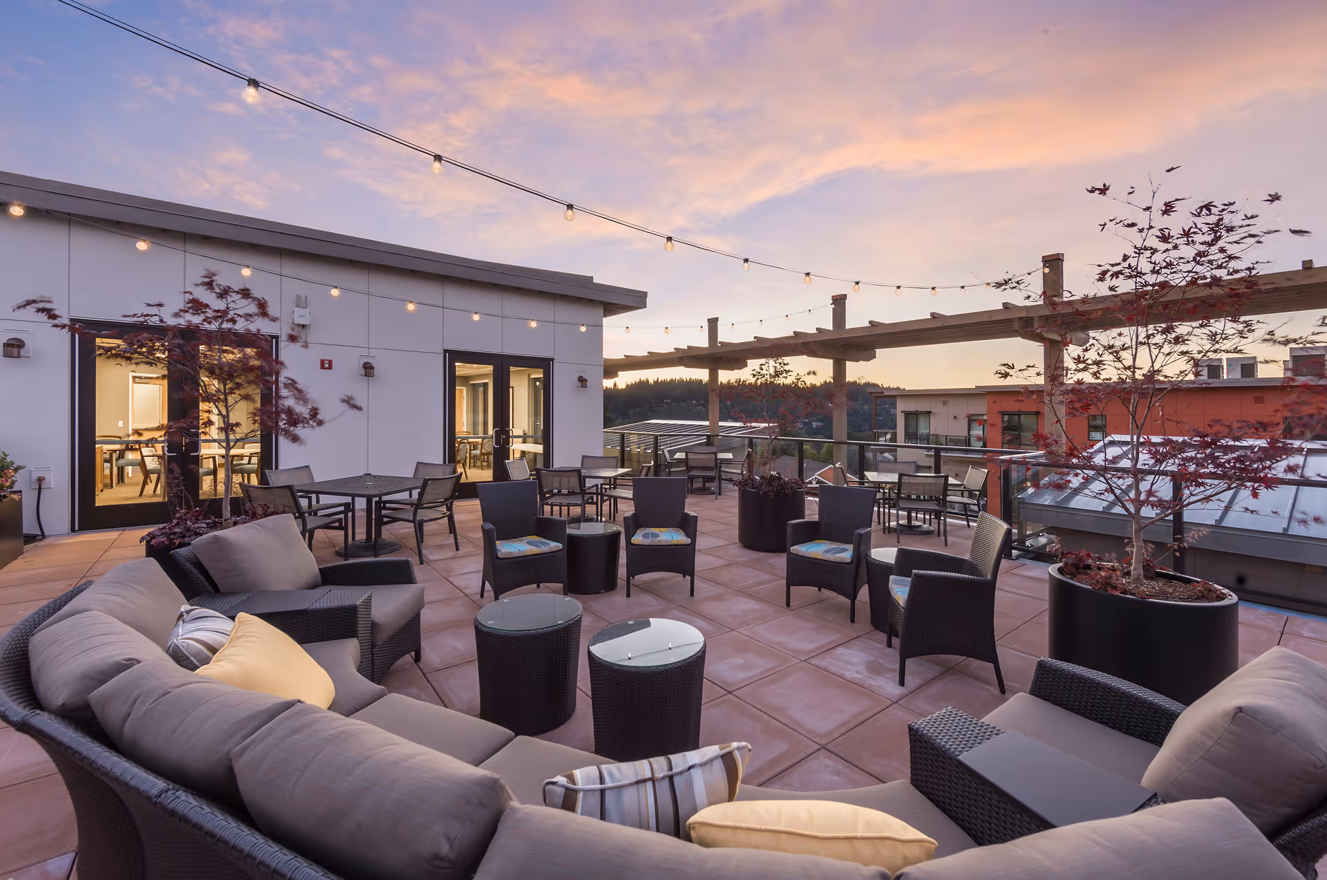 Outdoor patio area at sunset with cushioned seating, tables, chairs, potted plants, string lights overhead, and a pergola structure. The patio is part of a senior living community with glass doors leading inside.