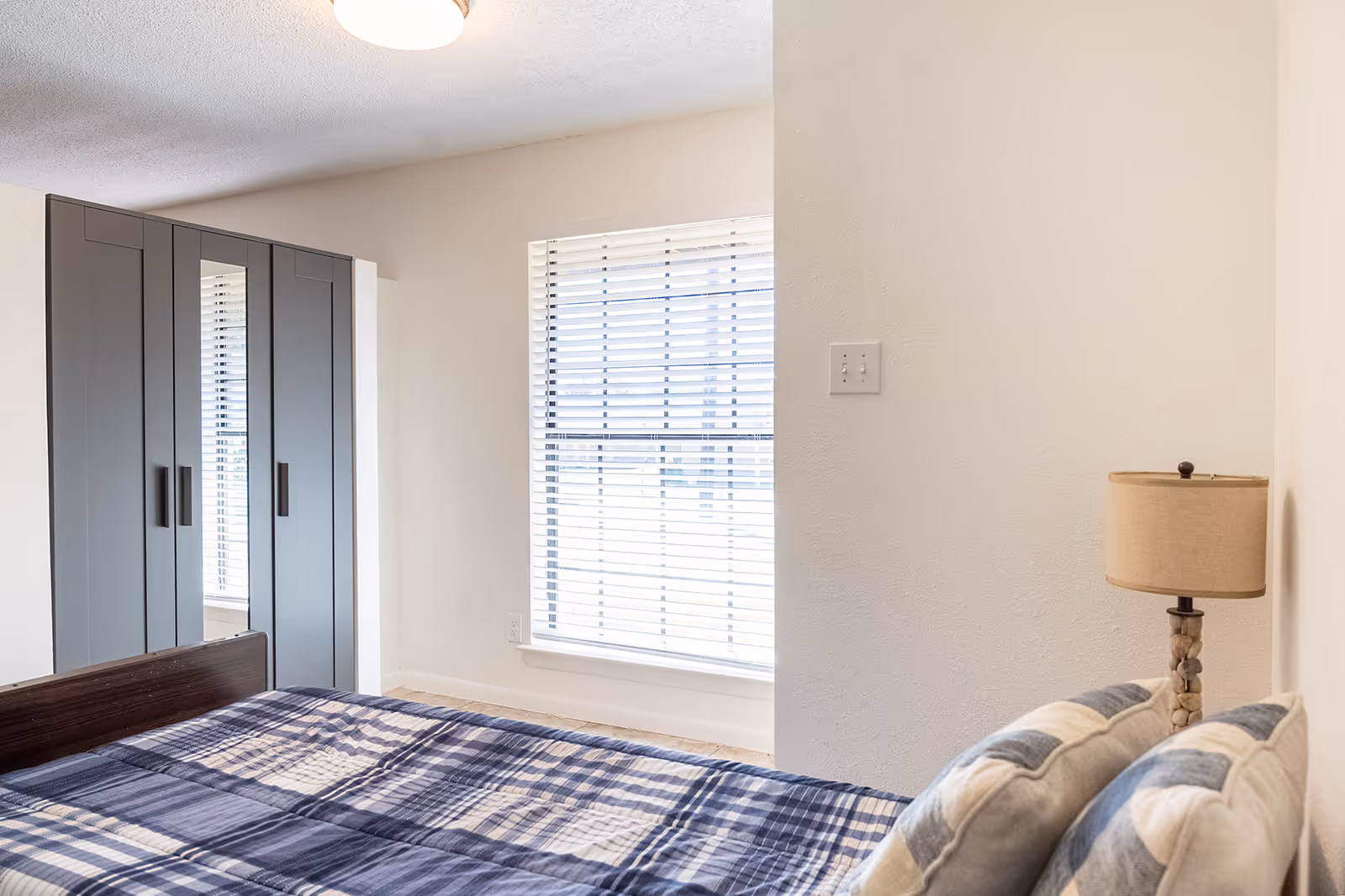 A bright bedroom with a bed covered in blue plaid bedding, pillows and a bedside lamp, a gray wardrobe and a window with blinds.