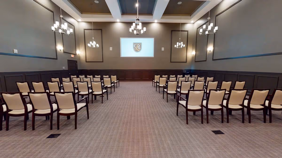 Empty meeting/event room with rows of beige chairs facing a projection screen and chandeliers.