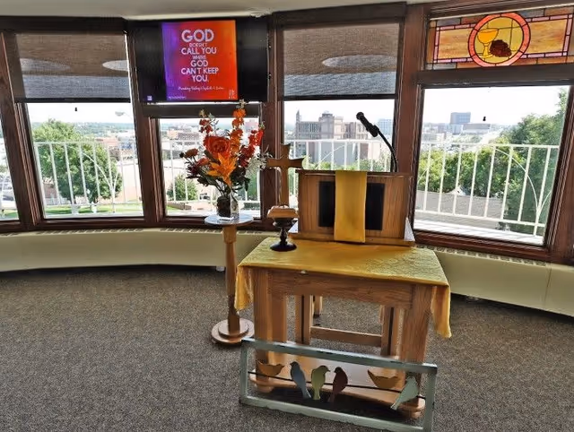 Small chapel-style interior with a wooden lectern draped in a yellow cloth, a microphone, vase of flowers, stained glass and large windows overlooking the city.