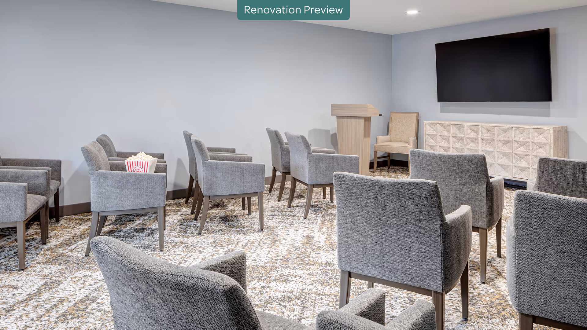 A small presentation or screening room with multiple gray upholstered chairs arranged in rows facing a large flat-screen TV mounted on the wall. There is a wooden podium and a single armchair near the TV, and a box of popcorn is placed on one of the chairs. The room has light-colored walls and a patterned carpet floor. A banner at the top reads 'Renovation Preview'.