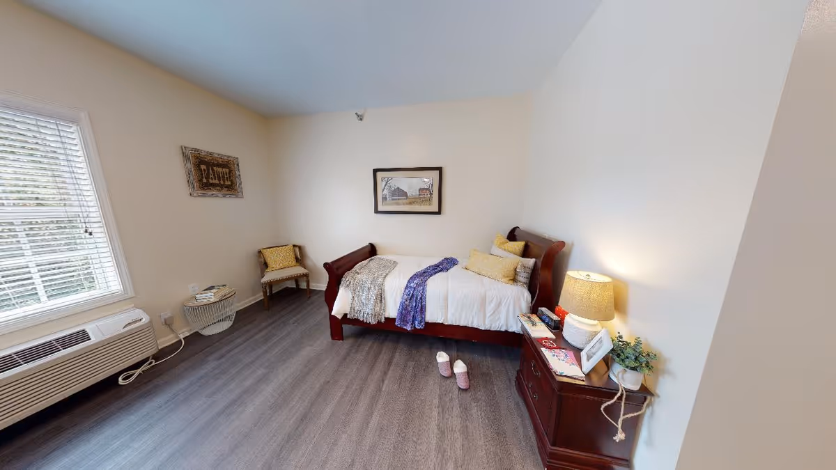 A cozy bedroom in a senior living facility featuring a wooden daybed with white bedding and multiple pillows. A purple and a beige throw blanket are draped over the bed. Next to the bed is a wooden nightstand with a lamp, framed photo, plant, and some books. A pair of pink slippers is placed on the floor beside the bed. There is a chair with a yellow cushion and a small round table with books near a window with white blinds. The walls are light-colored, and there is a framed picture and a decorative sign on the walls.