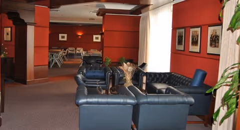 Interior view of a lounge area with black leather sofas arranged around a small table with a decorative centerpiece. The walls are painted red and adorned with framed pictures. In the background, there are tables and chairs set up, and large windows with sheer curtains allow natural light to enter the room.
