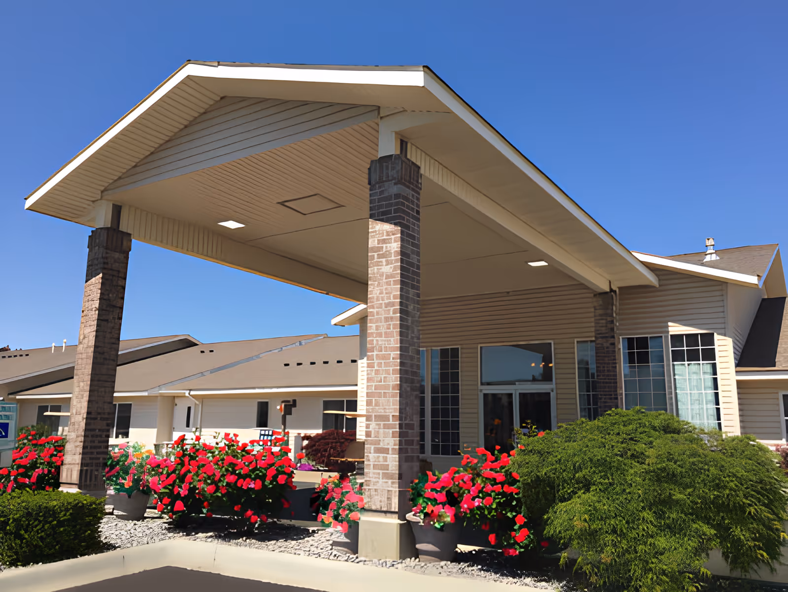 Exterior view of Green Lake Senior Living Amber Hills showing the entrance with a covered driveway supported by brick pillars, surrounded by vibrant red flowers and green shrubs under a clear blue sky.