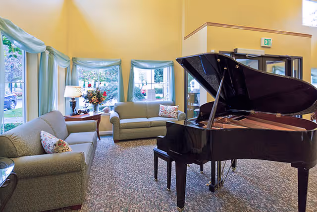A bright living room area with two light green sofas, a grand piano, and three windows with light blue curtains. There is a small table with a lamp and a flower arrangement between the sofas. The room has a patterned carpet and light yellow walls.