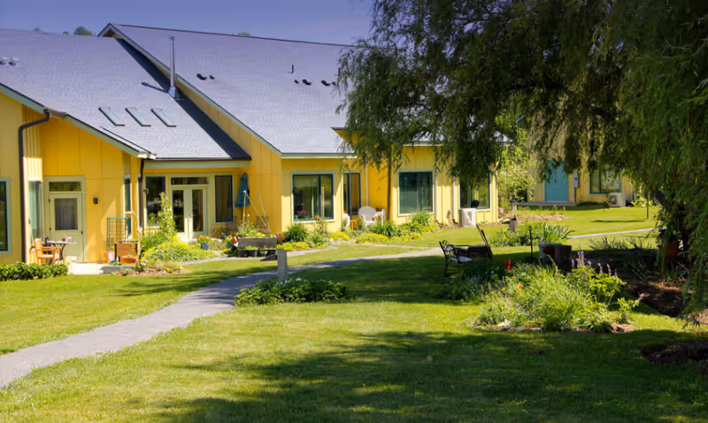 Yellow single-story building with a lawn, pathway, benches and gardens under a large tree.