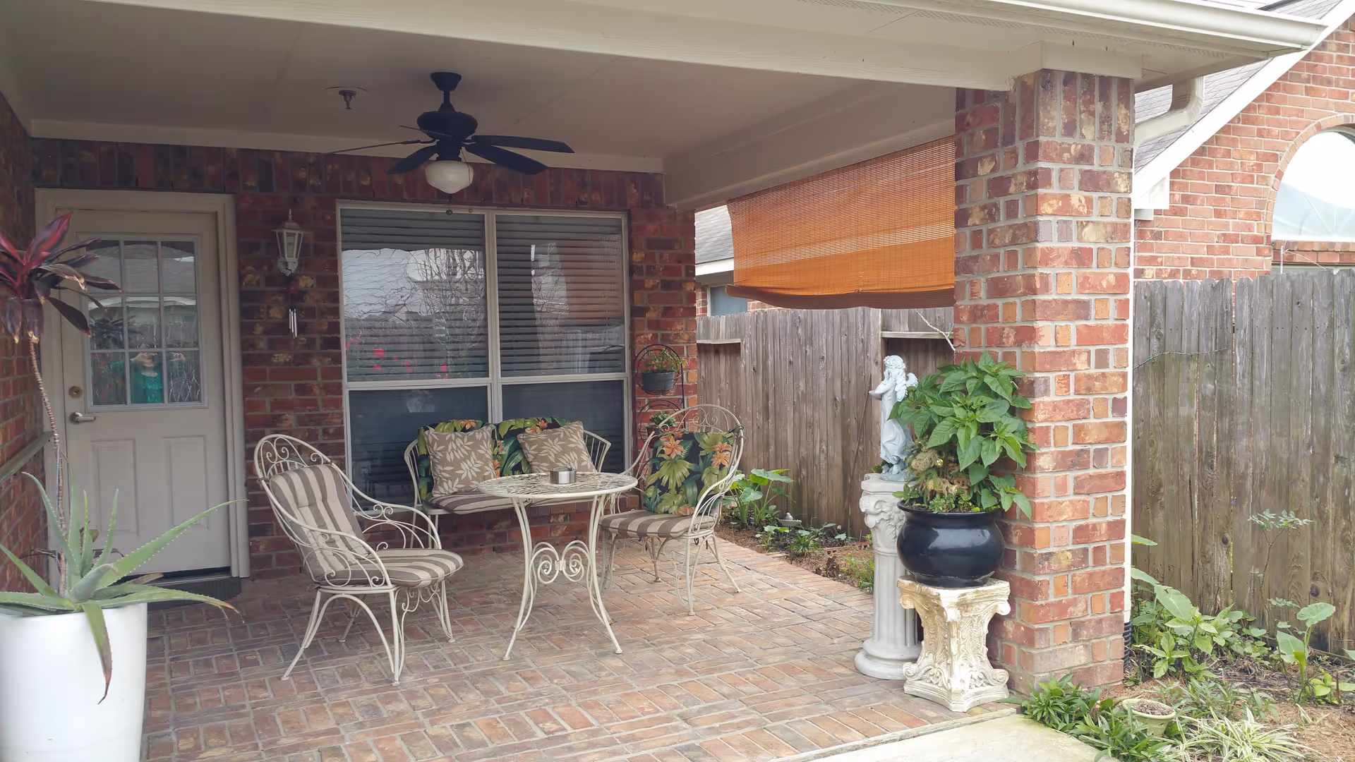 Covered outdoor patio area with brick flooring and brick pillars, featuring a white metal table and chairs with floral and striped cushions, potted plants, a ceiling fan, and a wooden fence in the background.