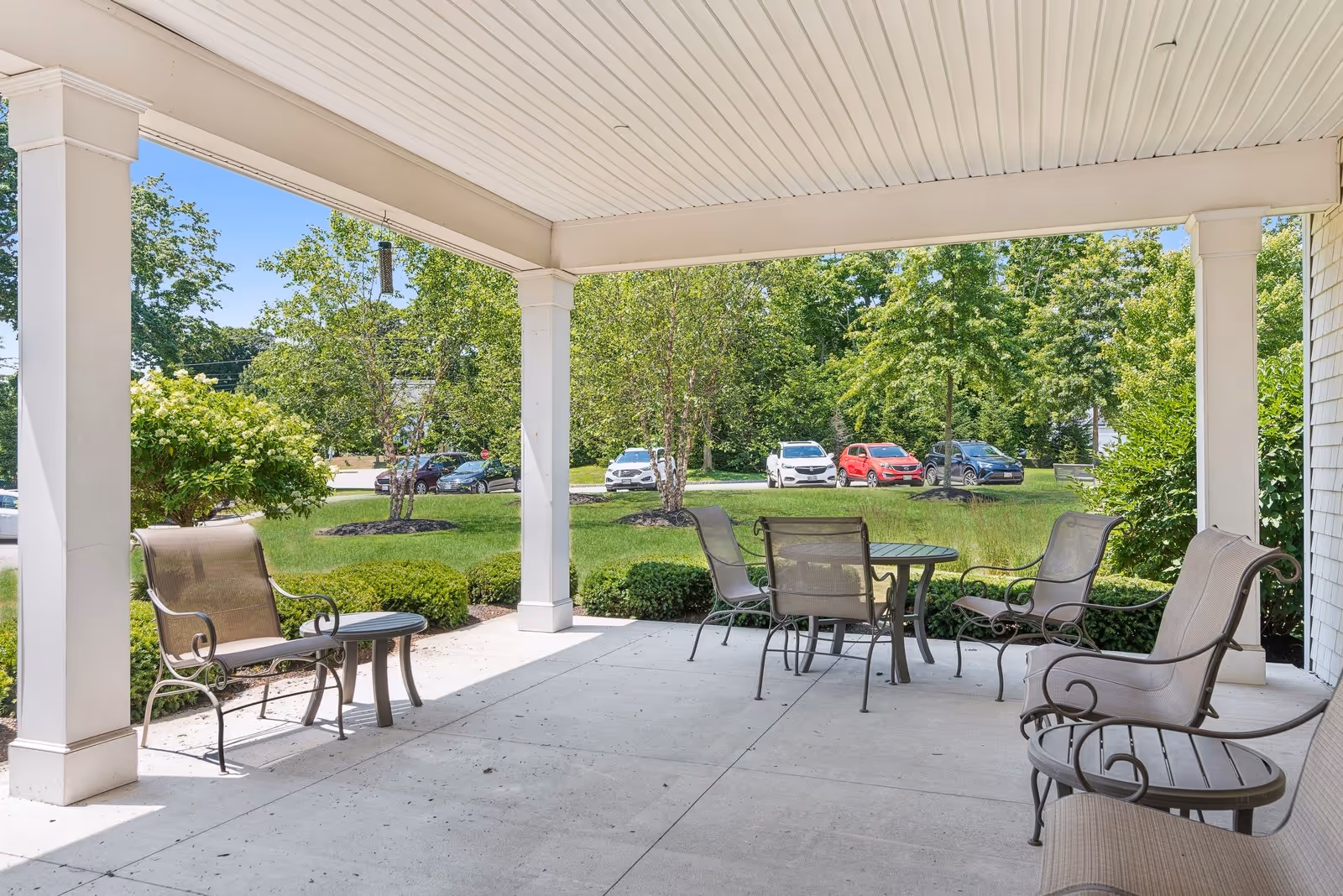 Covered outdoor patio area with metal chairs and tables, surrounded by green bushes and trees, with a parking lot and several cars visible in the background under a clear blue sky.