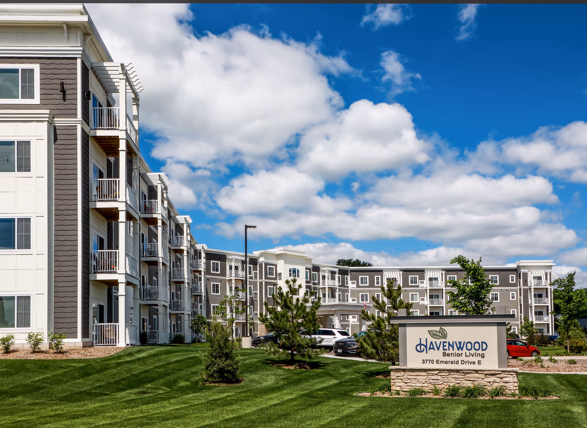 Exterior view of a multi-story senior living facility building with balconies, surrounded by green grass and small trees under a blue sky with scattered clouds. A sign in front reads 'Havenwood Senior Living, 3770 Emerald Drive E'.