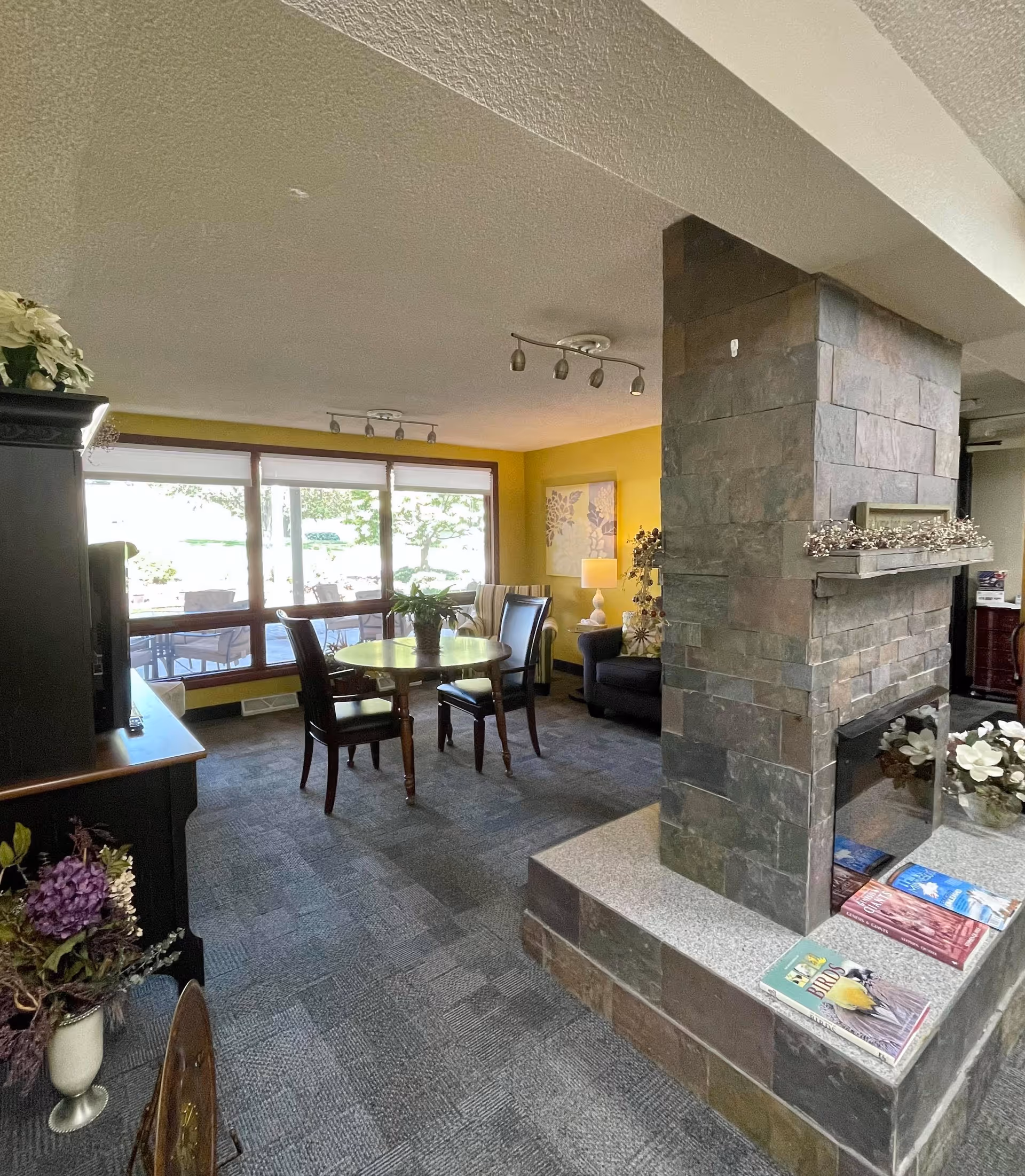 A cozy living room area with a stone fireplace in the center. There is a round wooden table with four chairs near large windows that let in natural light. The walls are painted yellow and decorated with artwork. A black armchair and a lamp are visible in the corner, along with some decorative plants and flowers.