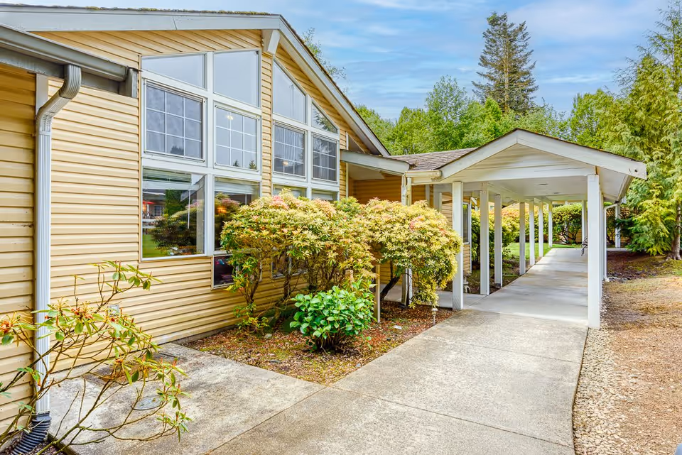 Covered walkway leading to the entrance of a yellow-sided building with large windows and landscaped shrubs.