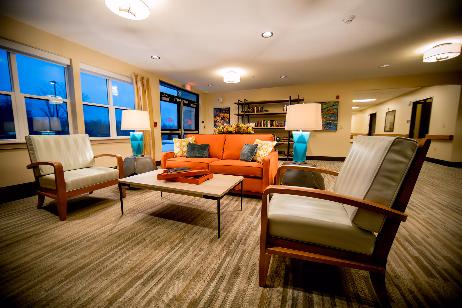 A cozy seating area in a senior living facility with an orange sofa adorned with decorative pillows, two cushioned armchairs with wooden frames, a rectangular coffee table with books and trays, two turquoise table lamps on side tables, a bookshelf with books and decor, and large windows showing an evening sky outside.
