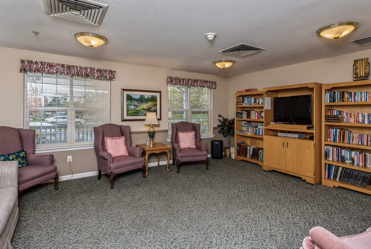 A cozy senior living common area with three purple armchairs, a wooden side table with a lamp, two large windows with floral valances, a painting on the wall, and wooden bookshelves filled with books and a TV.