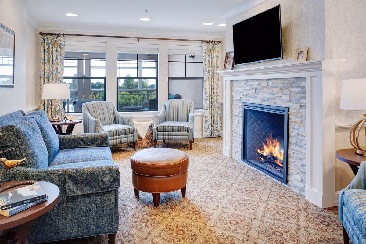 A cozy living room with a blue sofa, two striped armchairs, a round brown leather ottoman, and a fireplace with a fire burning. Above the fireplace is a mounted flat-screen TV. The room has large windows with floral curtains, patterned carpet, and two side tables with lamps.