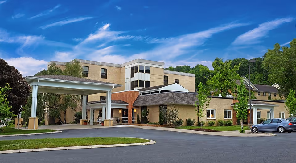 Exterior view of Cedarwood Rehabilitation and Healthcare Center showing a multi-story building with a covered entrance, surrounded by trees and a parking area with a car parked nearby under a blue sky with scattered clouds.