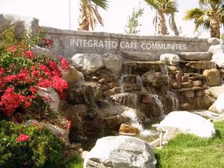A landscaped outdoor area featuring a stone waterfall with water cascading over rocks. The stone structure has the words 'INTEGRATED CARE COMMUNITIES' engraved on it. Surrounding the waterfall are green grass, bushes, and red flowering plants, with palm trees in the background.