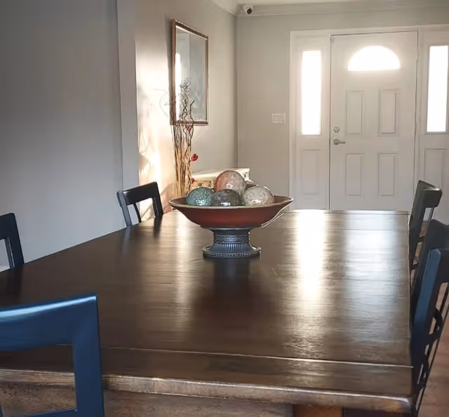 A dining room with a dark wooden table and black chairs. On the table is a decorative bowl filled with various ornamental spheres. In the background, there is a white front door with sidelights and a mirror on the wall reflecting some light.