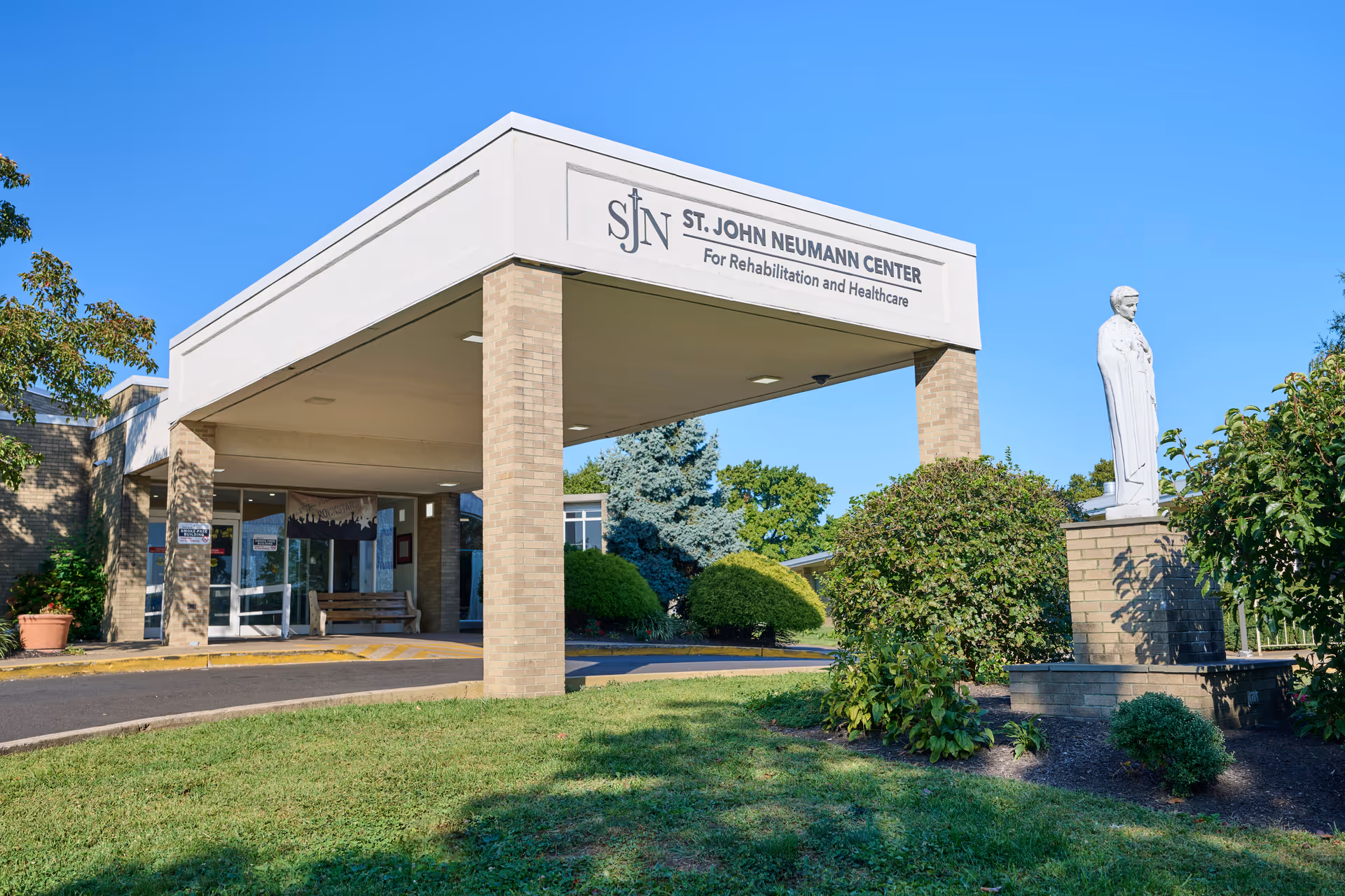 Exterior view of the St. John Neumann Center for Rehabilitation and Healthcare building entrance with a covered drop-off area supported by brick pillars. There is a statue on a brick pedestal surrounded by bushes and greenery under a clear blue sky.