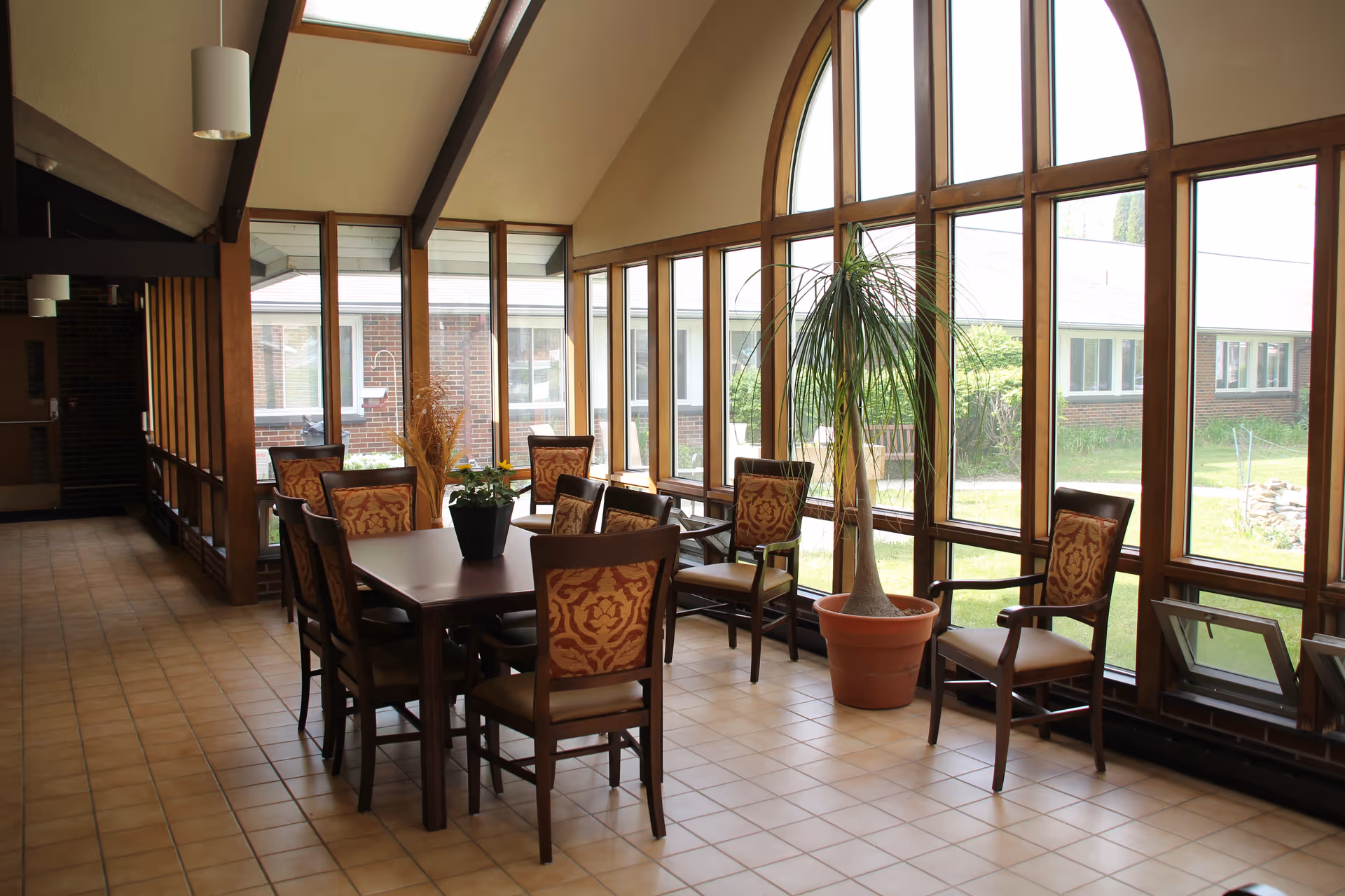 A sunlit interior room with large arched windows along one wall, featuring a wooden table surrounded by eight upholstered chairs with patterned backs. There is a potted plant on the table and a tall potted plant near the windows. The floor is tiled and the ceiling has exposed beams with hanging light fixtures.