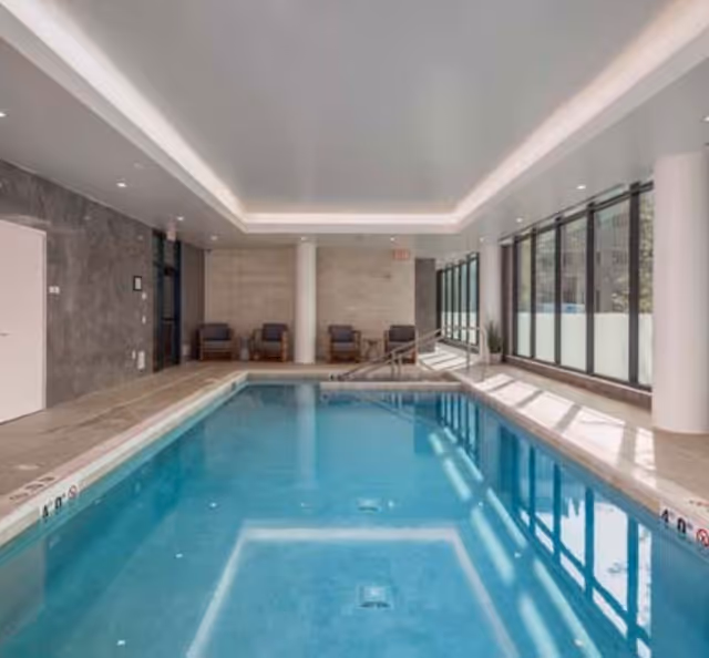 Indoor swimming pool with lounge chairs, floor-to-ceiling windows, and recessed ceiling lighting.