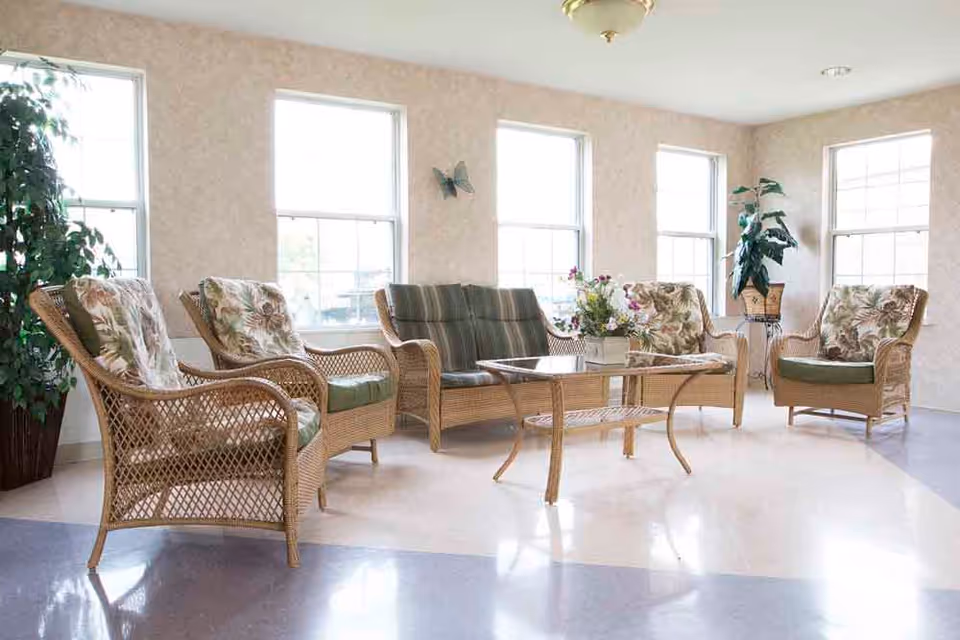 A bright sitting area with wicker furniture including four armchairs and a loveseat, all with floral and striped cushions. A glass-top wicker coffee table with a flower arrangement is in the center. The room has several large windows letting in natural light and two potted plants in the corners.