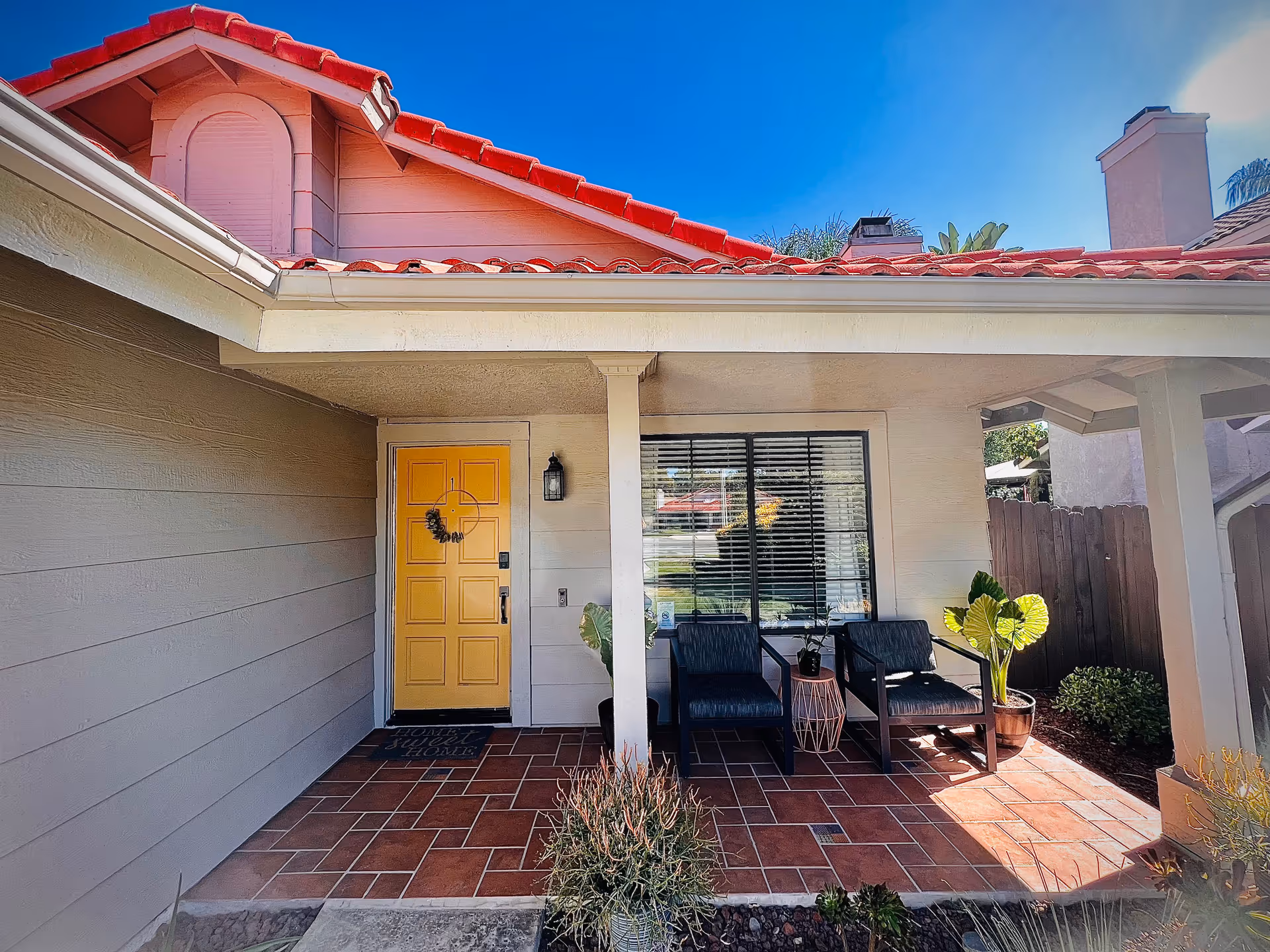 Front porch of a house with a yellow door, two dark blue chairs, a small table with a plant, and potted plants on a tiled floor under a covered roof with red tiles.